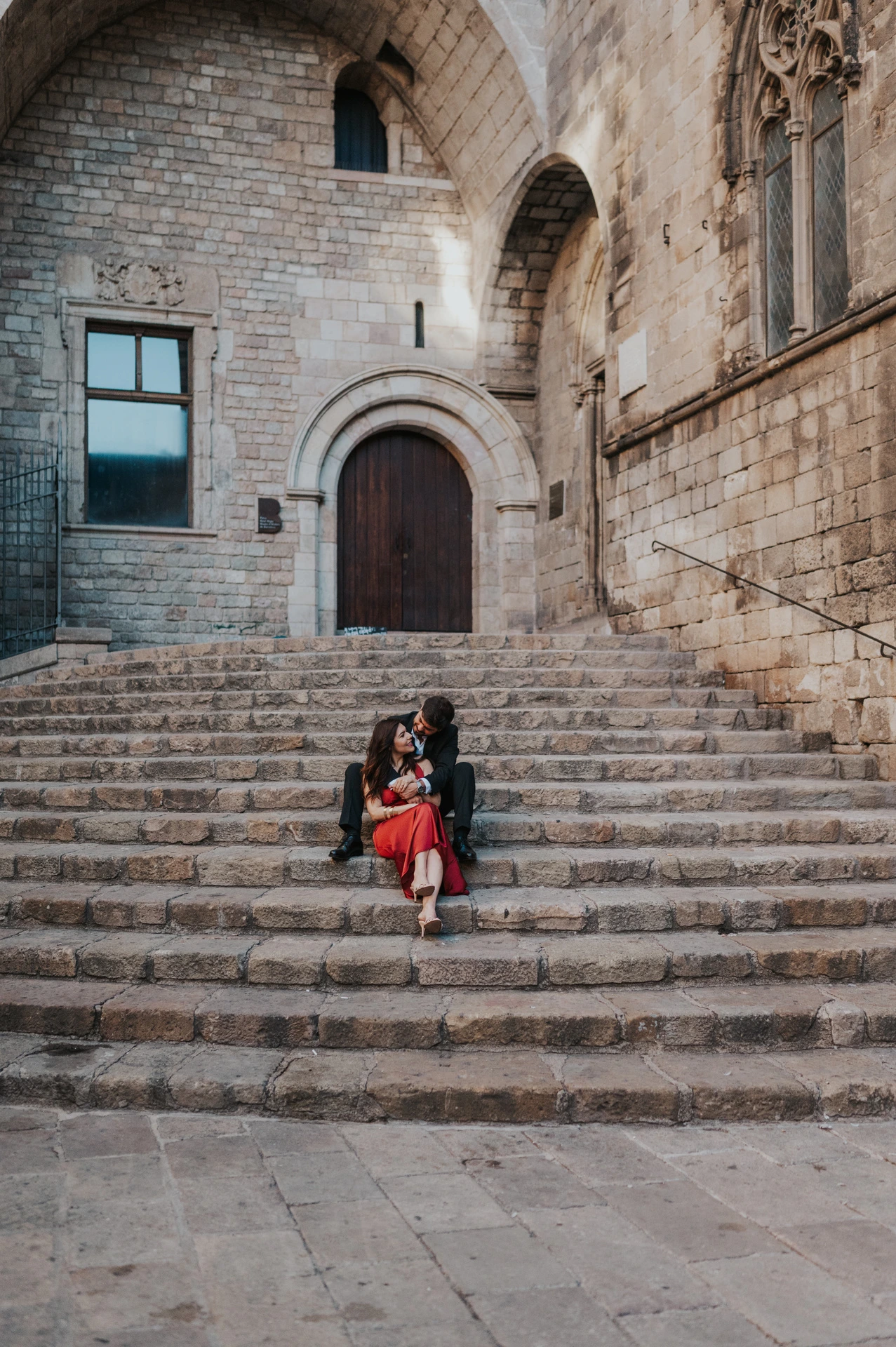 The couple sitting close together on the stone steps at Plaça del Rei, beneath an arched doorway during their engagement photoshoot in Barcelona.