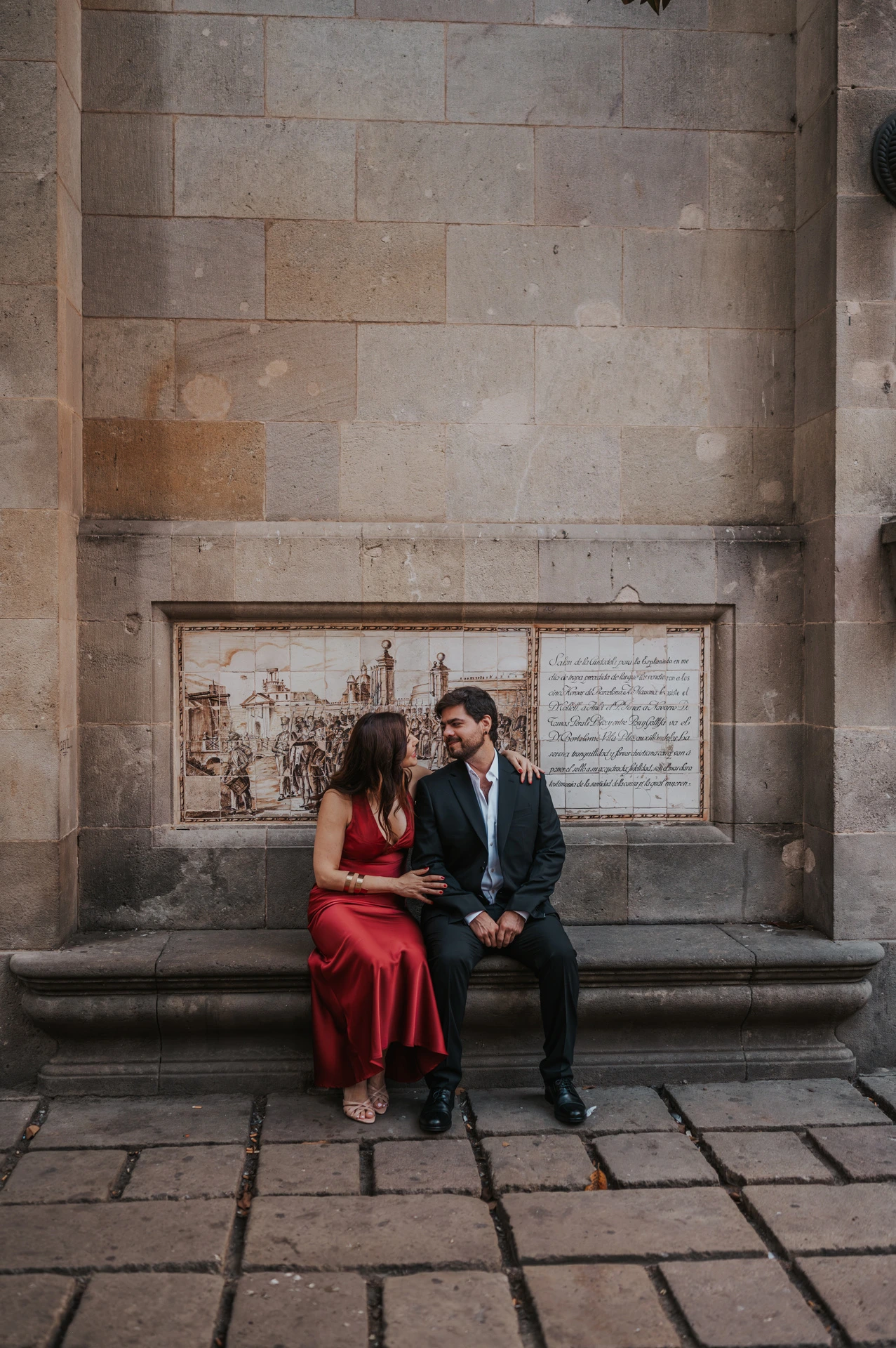 The couple sitting together on a stone bench against a tall historic wall in the Gothic Quarter during their engagement photoshoot in Barcelona.