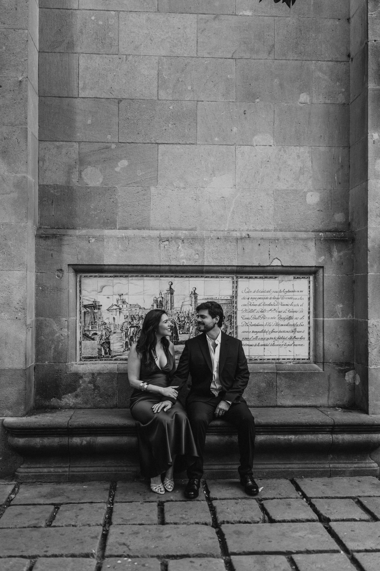 Black-and-white photograph: the couple sitting together on a stone bench against a tall historic wall in the Gothic Quarter during their engagement photoshoot in Barcelona.