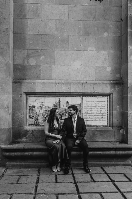 Together on a stone bench in the old town in black and white | Barcelona engagement photoshoot Black-and-white photograph: the couple sitting together on a stone bench against a tall historic wall in the Gothic Quarter during their engagement photoshoot in Barcelona.