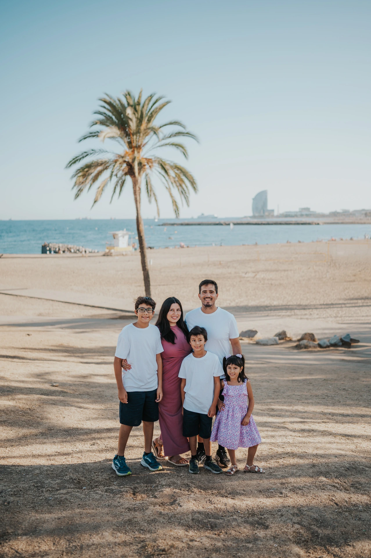 The parents and their three children standing together beneath a palm tree, with the beach and W Hotel behind them during their family photoshoot in Barcelona.