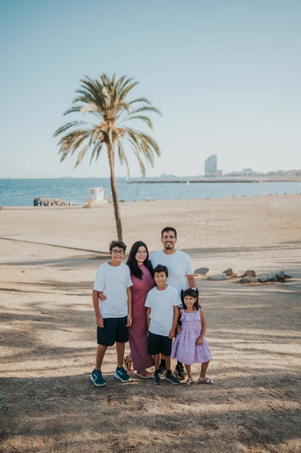 Family portraits beneath a beach palm tree | Barcelona family photoshoot The parents and their three children standing together beneath a palm tree, with the beach and W Hotel behind them during their family photoshoot in Barcelona.