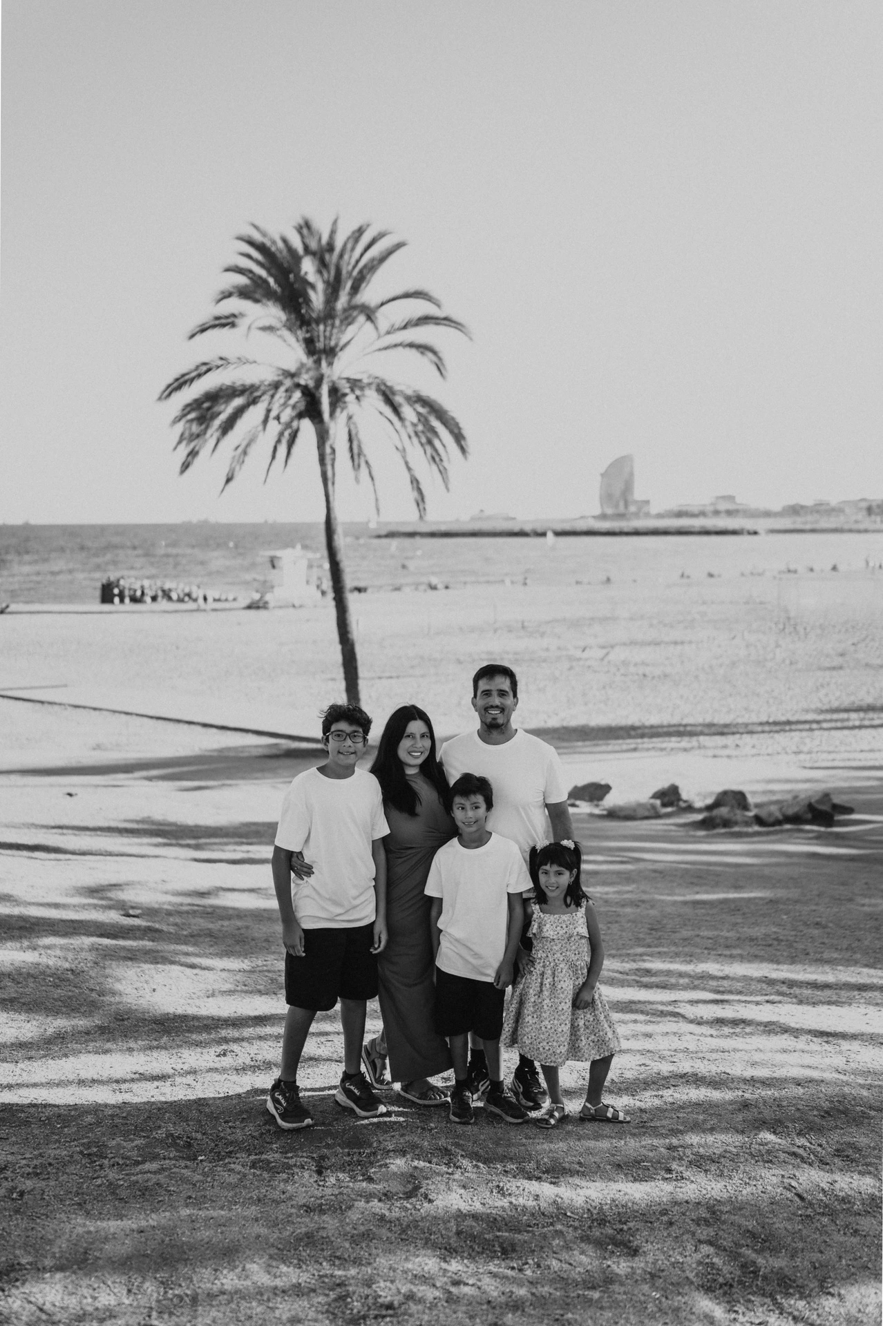 Black-and-white photograph of the parents and their three children standing together beneath a palm tree, with the beach and W Hotel behind them during their family photoshoot in Barcelona.