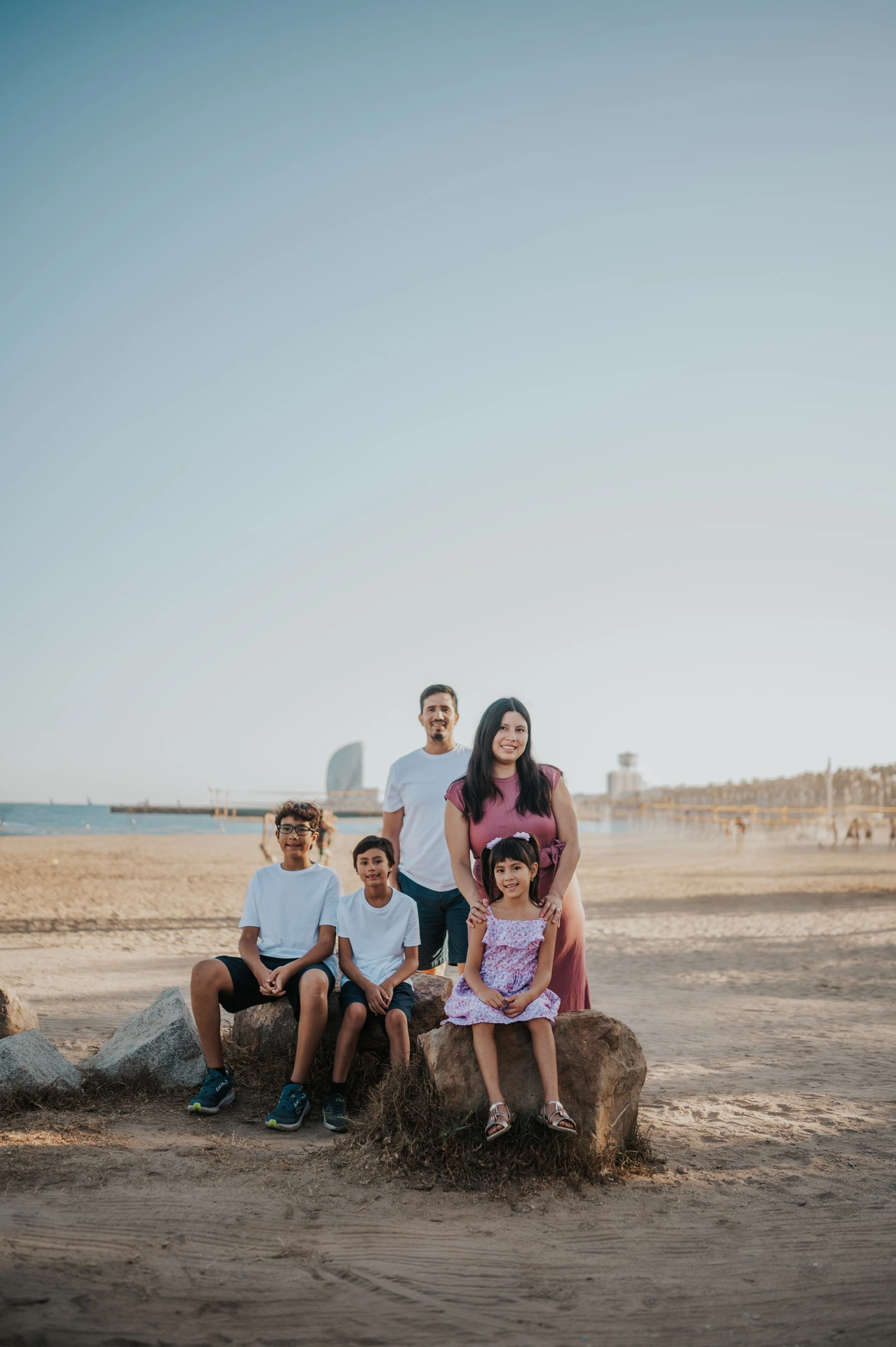 The family sitting and standing together on rocks beside the beach, with the W Hotel across the water during their family photoshoot in Barcelona.