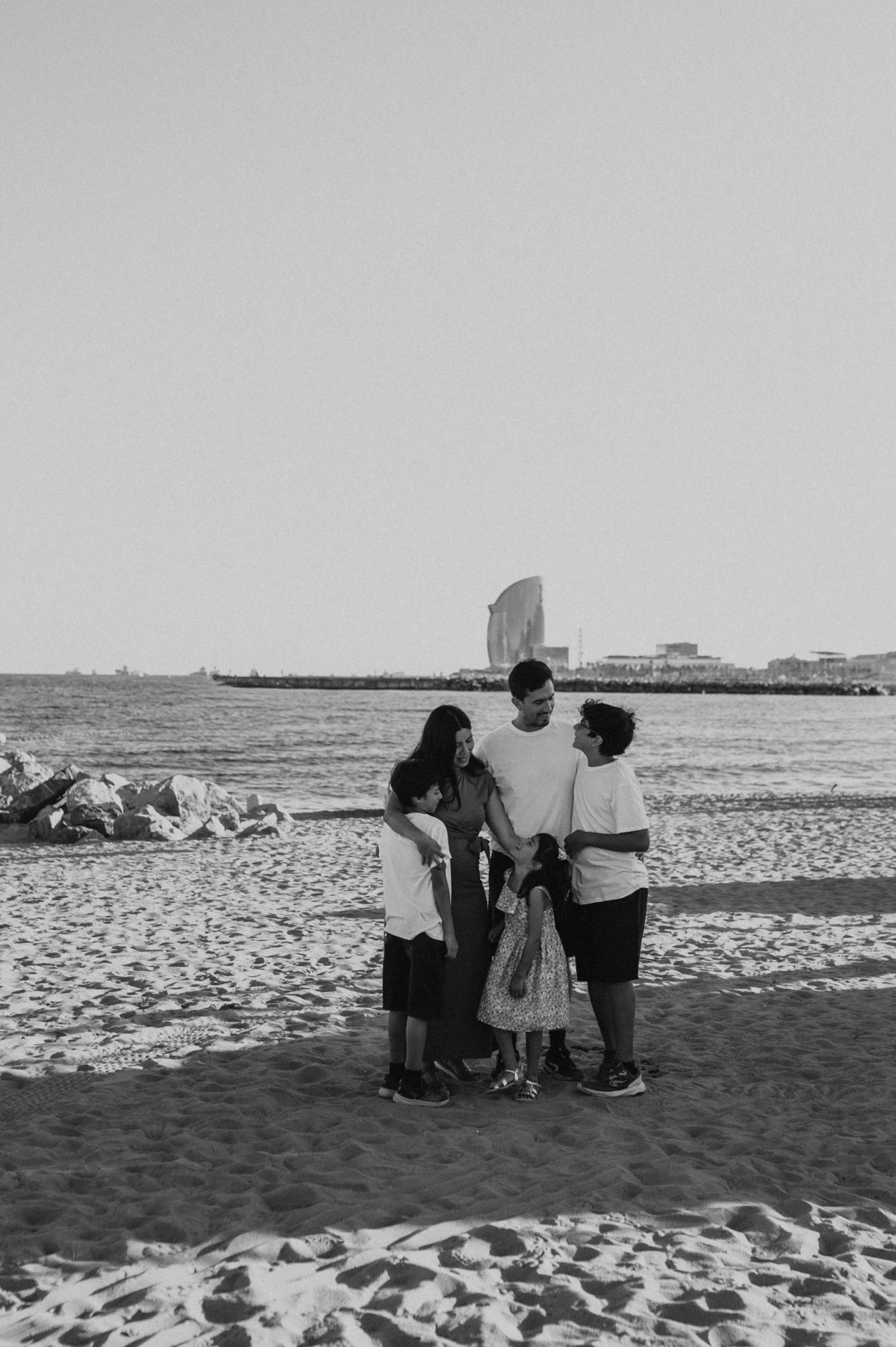 Black-and-white photograph of the parents and their three children gathered closely together on the sand beside the sea during their family photoshoot in Barcelona.
