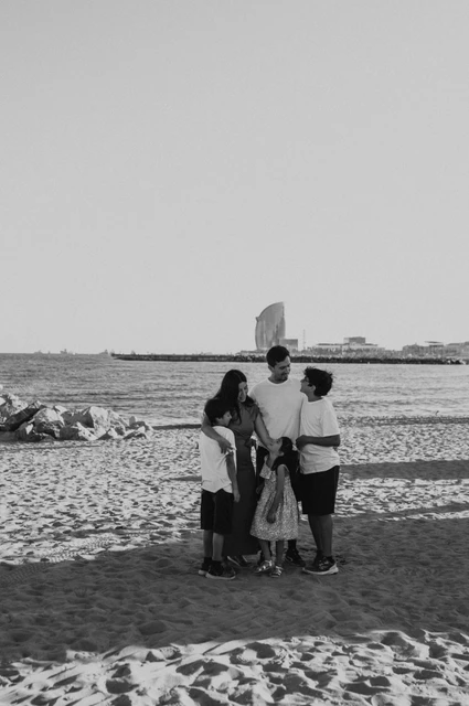Together by the sea on the sand in black and white | Barcelona family photoshoot Black-and-white photograph of the parents and their three children gathered closely together on the sand beside the sea during their family photoshoot in Barcelona.