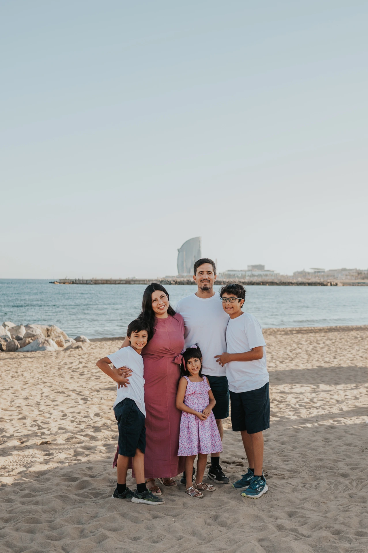 The family of five standing close together on the sand with the sea and W Hotel behind them during their family photoshoot in Barcelona.