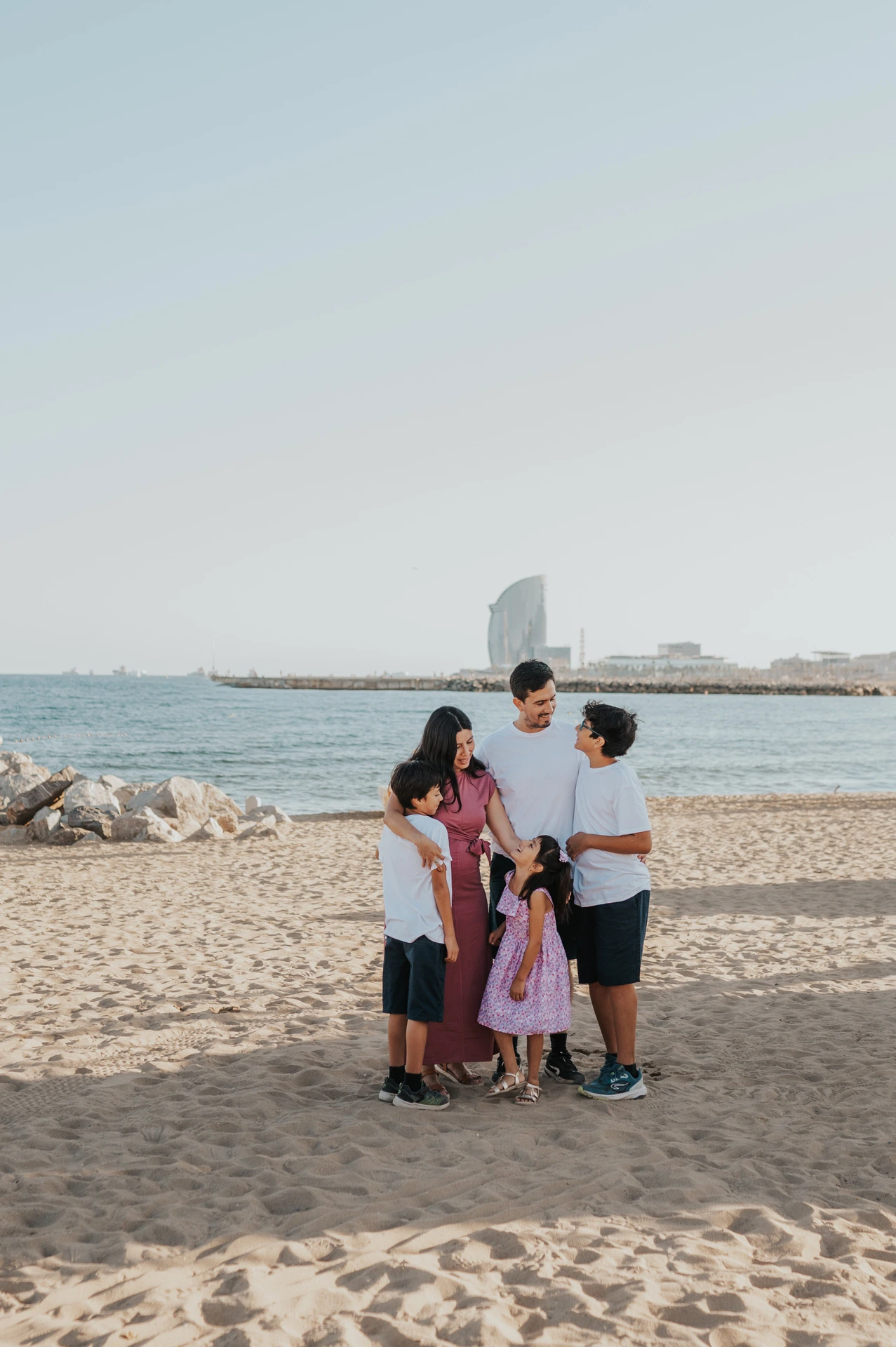 The parents and their three children gathered closely together on the sand beside the sea during their family photoshoot in Barcelona.