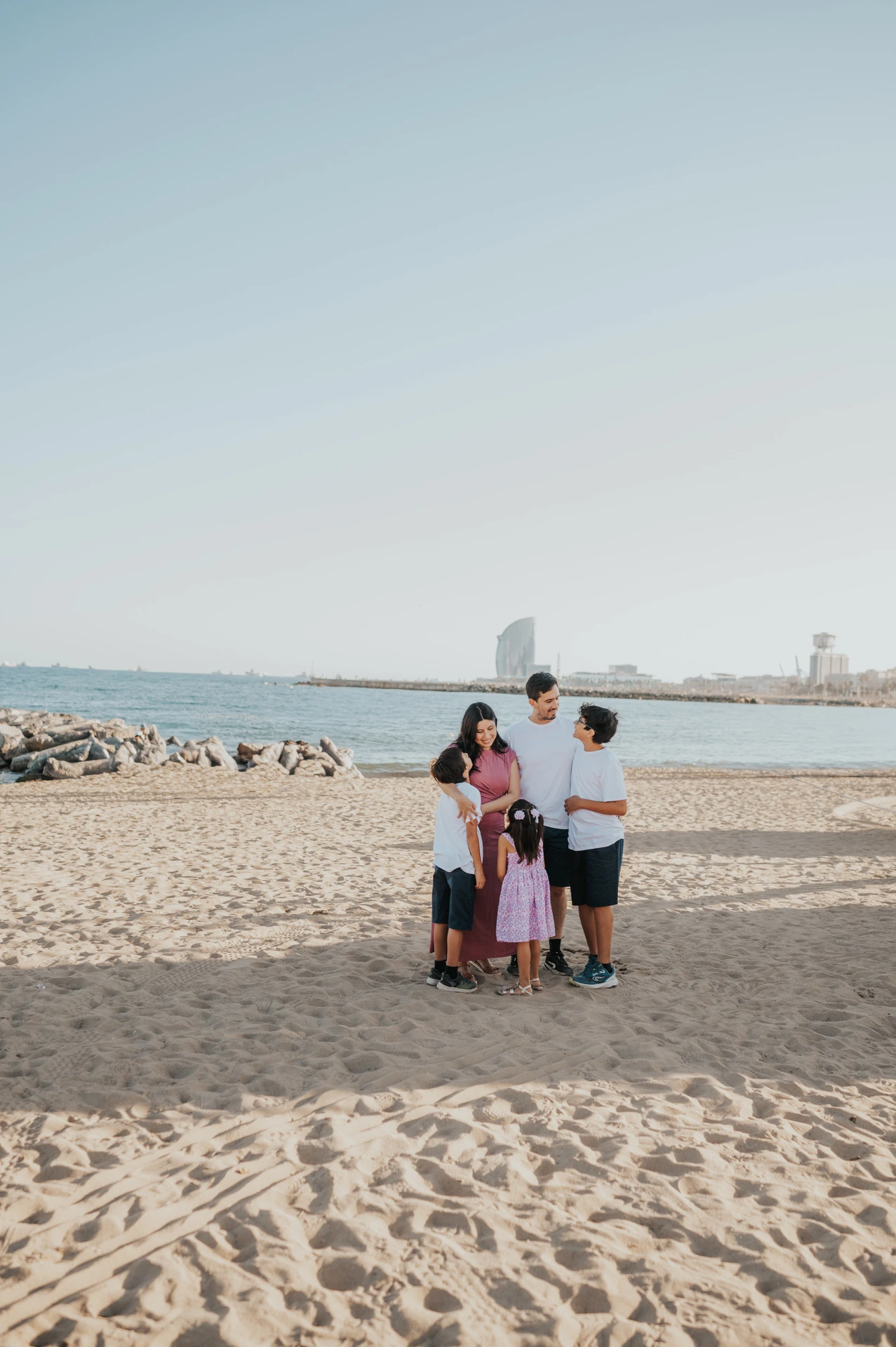 A wide view of the family gathered together on the sand beside the sea, with the W Hotel in the distance during their family photoshoot in Barcelona.