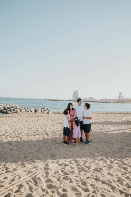 A wide seaside family portrait | Barcelona family photoshoot A wide view of the family gathered together on the sand beside the sea, with the W Hotel in the distance during their family photoshoot in Barcelona.