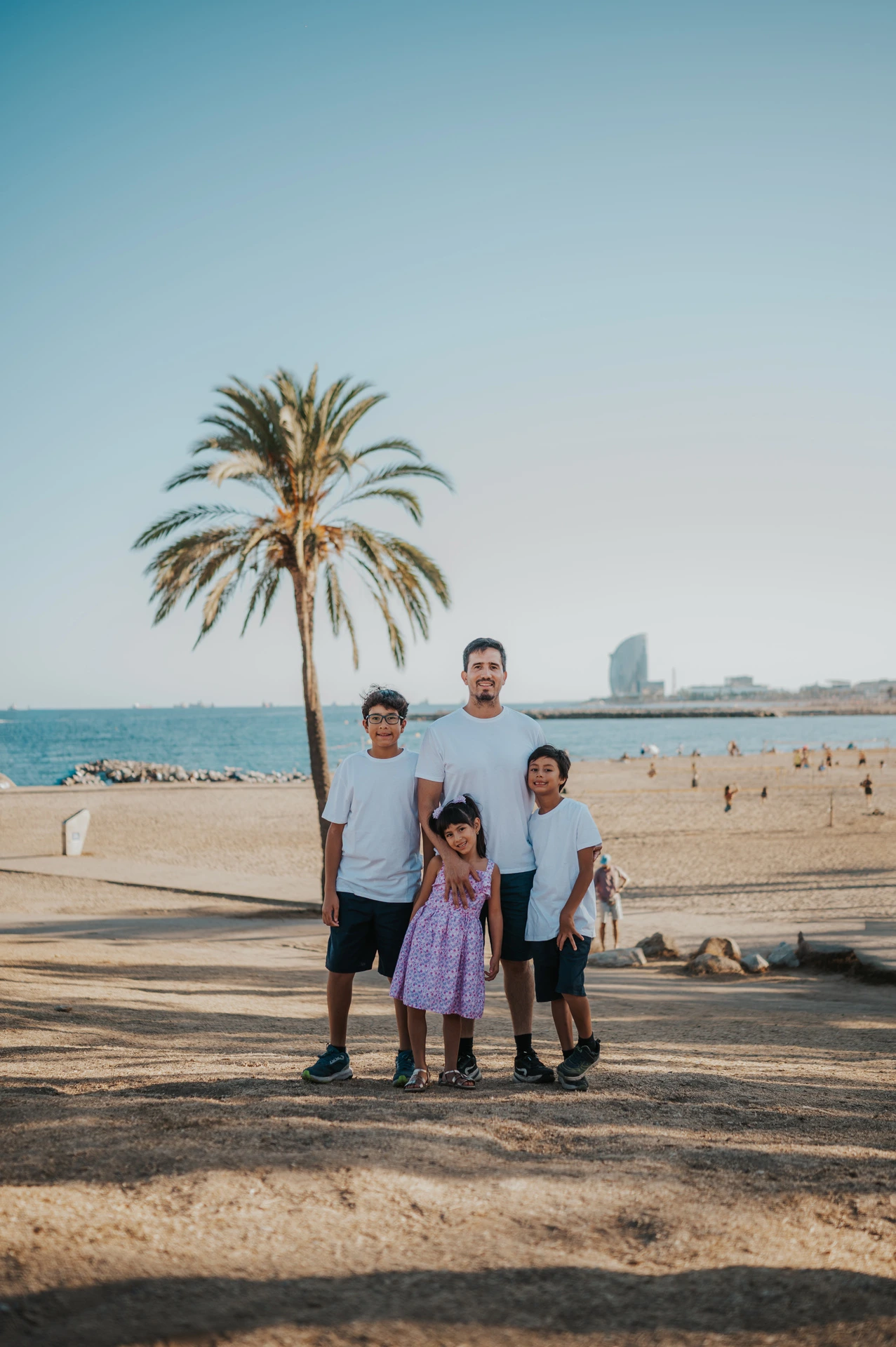 The father standing with his three children beside a palm tree overlooking the beach during their family photoshoot in Barcelona.
