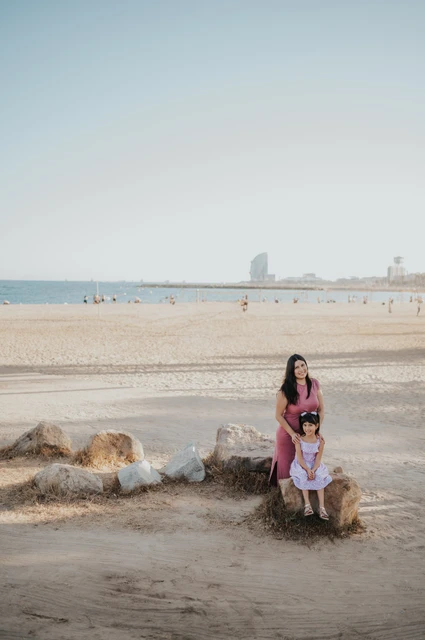 Mother and daughter on the beachside rocks | Barcelona family photoshoot The mother standing behind her daughter, who is sitting on a beachside rock, with the sea and W Hotel in the distance during their family photoshoot in Barcelona.
