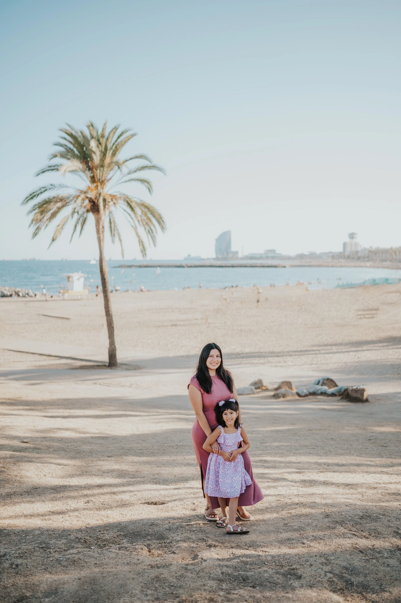 The mother and daughter standing together beside a palm tree overlooking the beach and sea during their family photoshoot in Barcelona.