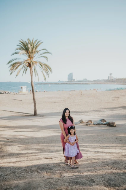 The mother and daughter standing together beside a palm tree overlooking the beach and sea during their family photoshoot in Barcelona.