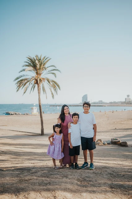 A mother and her three children by the beach | Barcelona family photoshoot The mother standing with her three children beneath a palm tree beside the beach during their family photoshoot in Barcelona.