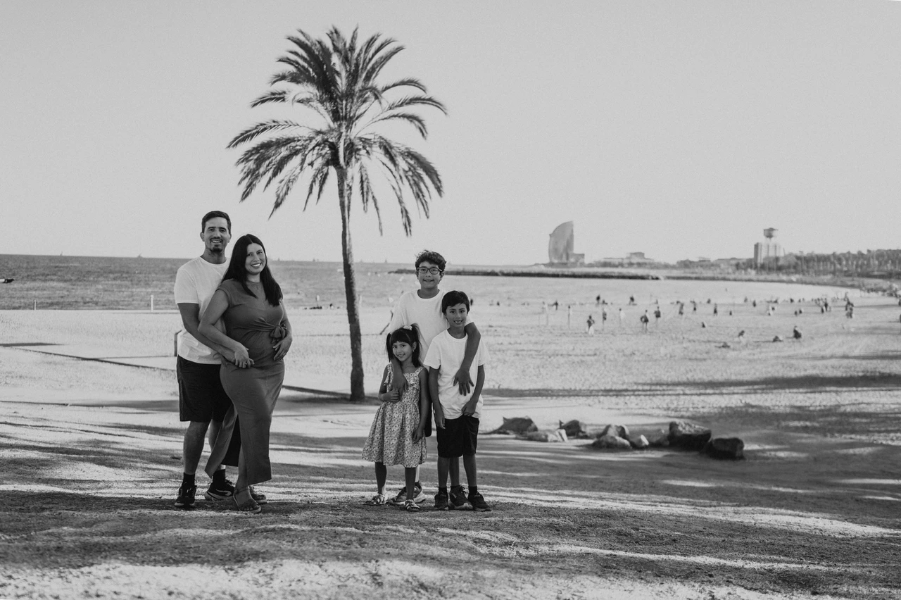 Parents and children with a sunny beach backdrop in black and white | Barcelona family photoshoot Black-and-white photograph of the parents standing together beside a palm tree while their three children stand nearby, with the beach and W Hotel behind them during their family photoshoot in Barcelona.