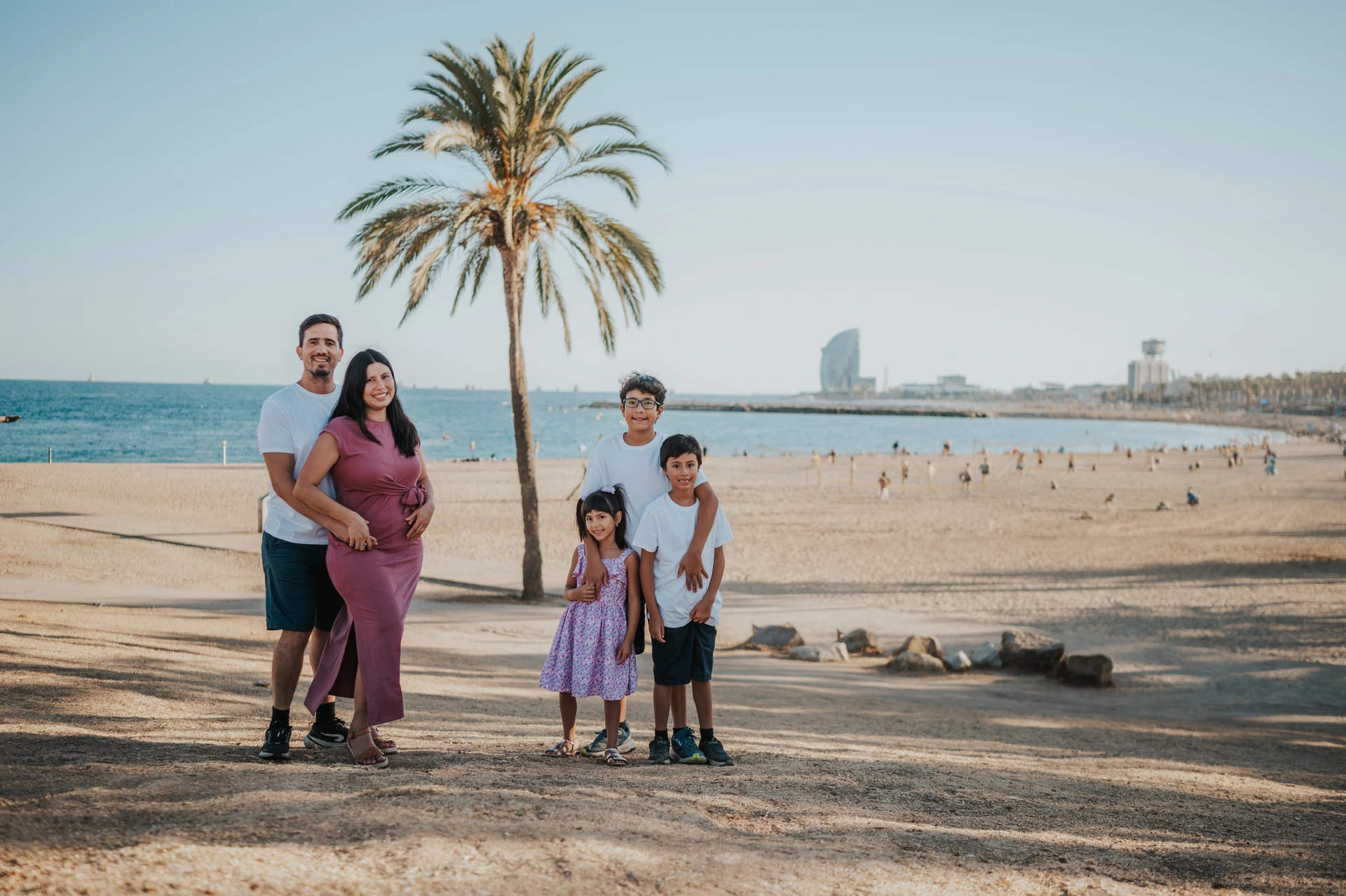 Parents and children with a sunny beach backdrop | Barcelona family photoshoot The parents standing together beside a palm tree while their three children stand nearby, with the beach and W Hotel behind them during their family photoshoot in Barcelona.