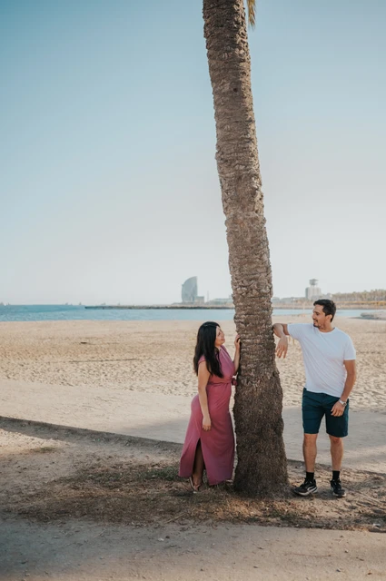 The parents standing on opposite sides of a palm tree and looking towards one another beside the beach during their family photoshoot in Barcelona.