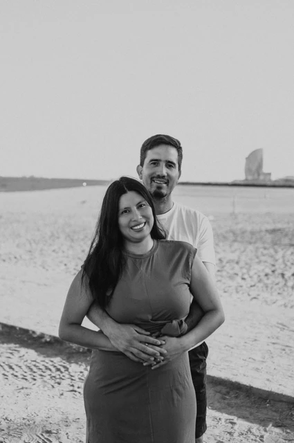 Black-and-white photograph of the parents embracing on the sand, with the sea and W Hotel behind them during their family photoshoot in Barcelona.