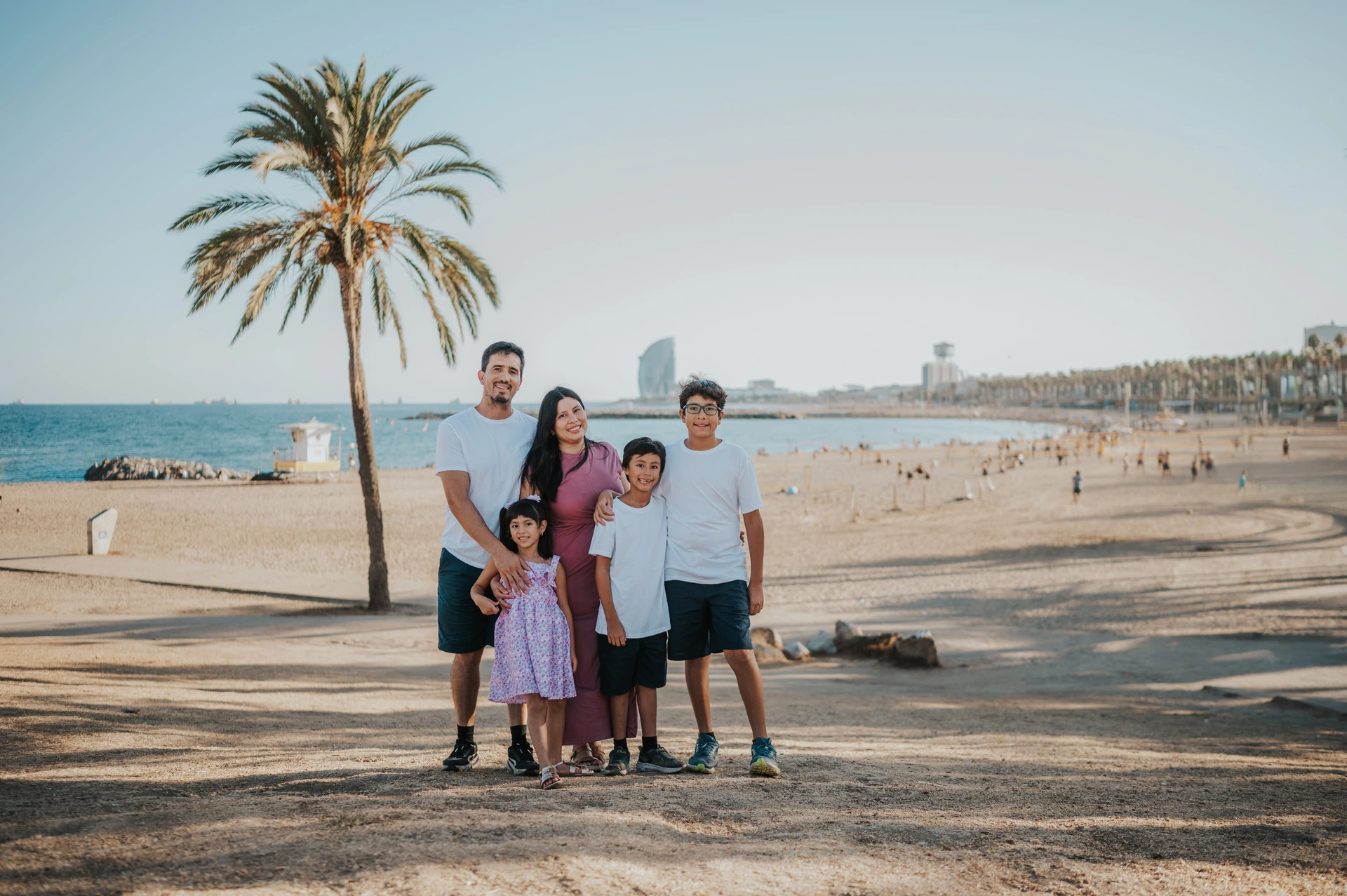 The whole family with the beach and W Hotel behind them | Barcelona family photoshoot The family of five standing together beside a palm tree overlooking the beach, sea and W Hotel during their family photoshoot in Barcelona.