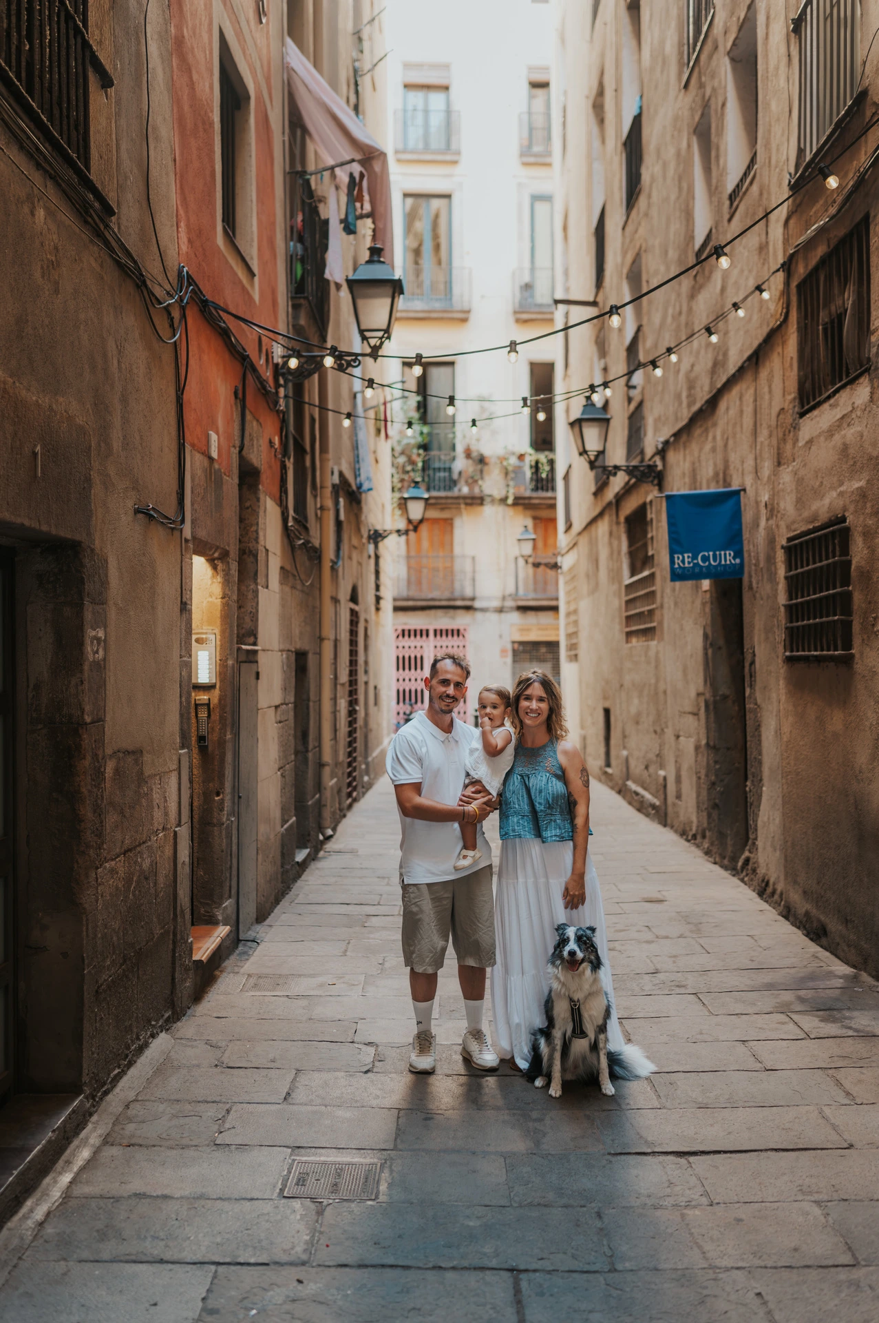 The parents holding their toddler with their dog sitting beside them on an El Born street during their family photoshoot in Barcelona.