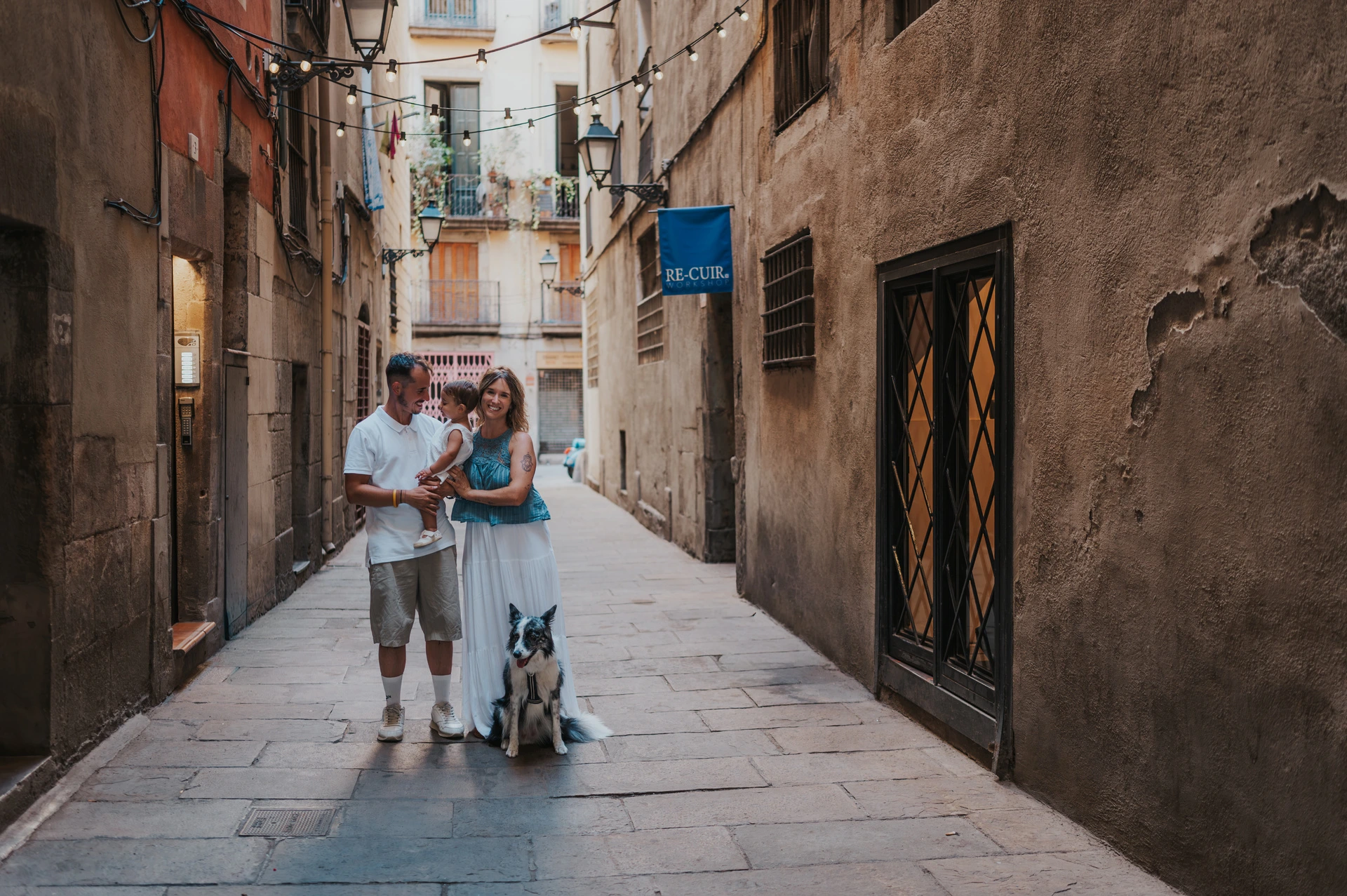 The parents holding their toddler with their dog sitting beside them on an El Born street during their family photoshoot in Barcelona.