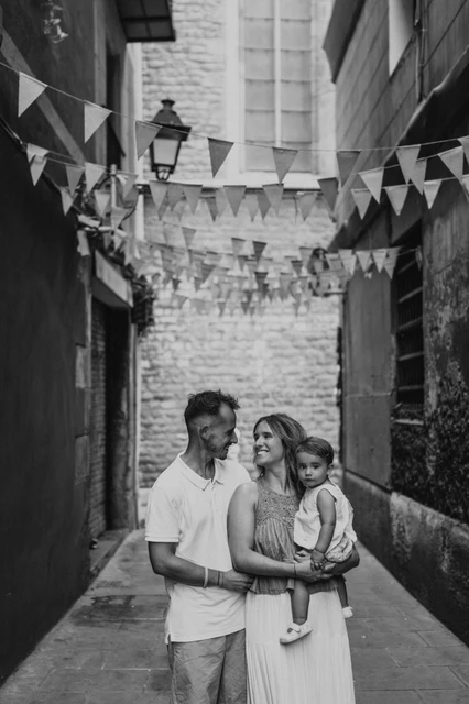 Family portraits beneath colourful bunting in El Born in black and white | Barcelona family photoshoot Black-and-white photograph: the parents standing close together with their toddler beneath colourful bunting in an El Born lane during their family photoshoot in Barcelona.