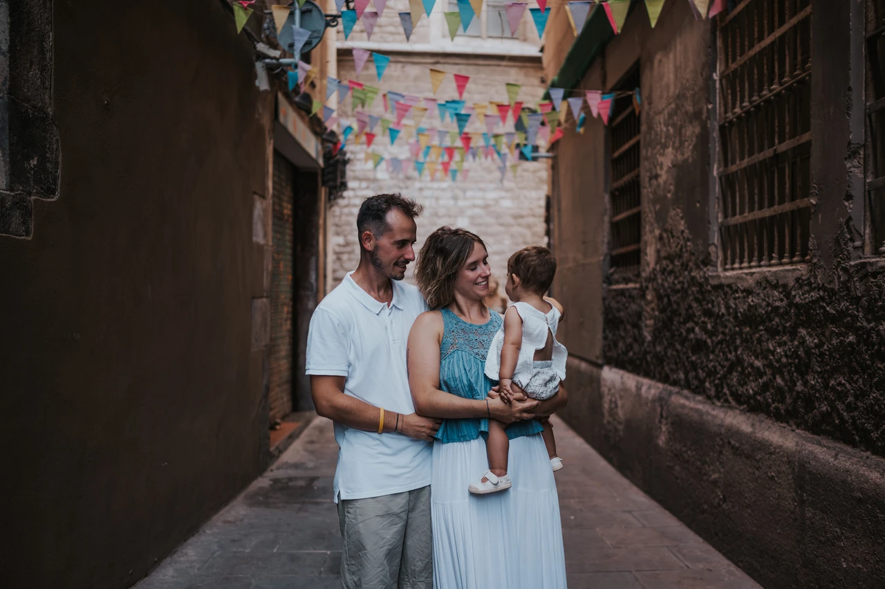 Family portraits beneath colourful bunting in El Born | Barcelona family photoshoot The parents standing close together with their toddler beneath colourful bunting in an El Born lane during their family photoshoot in Barcelona.