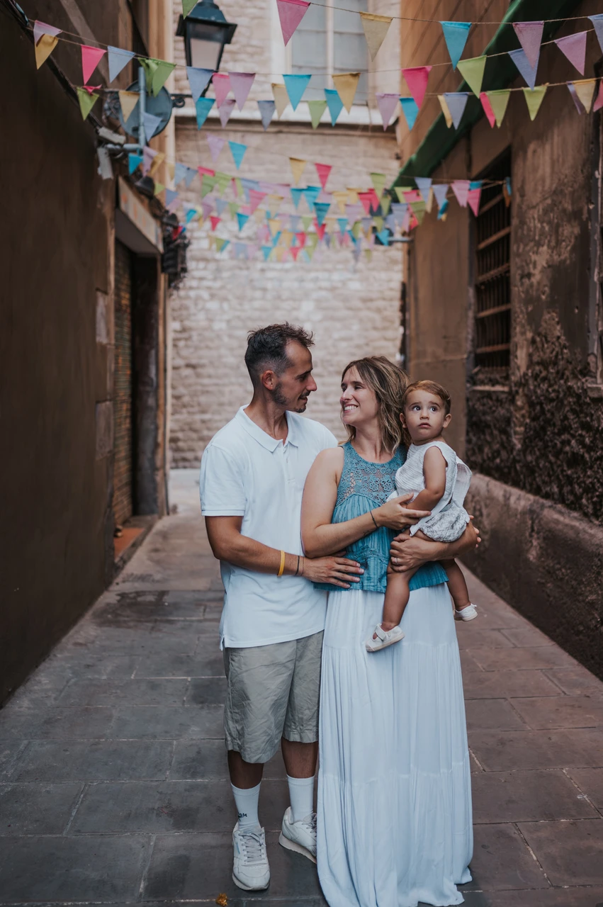 Family portraits beneath colourful bunting in El Born | Barcelona family photoshoot The parents standing close together with their toddler beneath colourful bunting in an El Born lane during their family photoshoot in Barcelona.