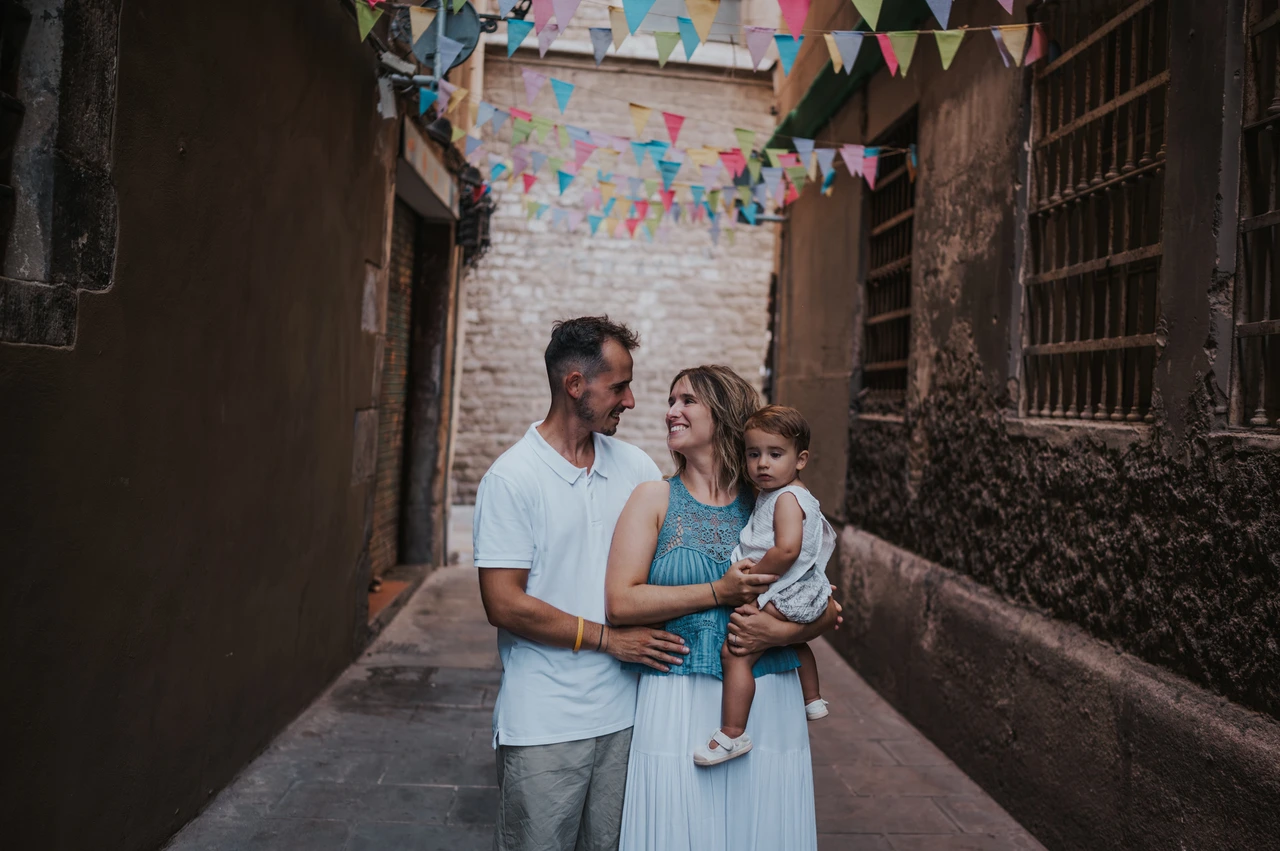 Family portraits beneath colourful bunting in El Born | Barcelona family photoshoot The parents standing close together with their toddler beneath colourful bunting in an El Born lane during their family photoshoot in Barcelona.