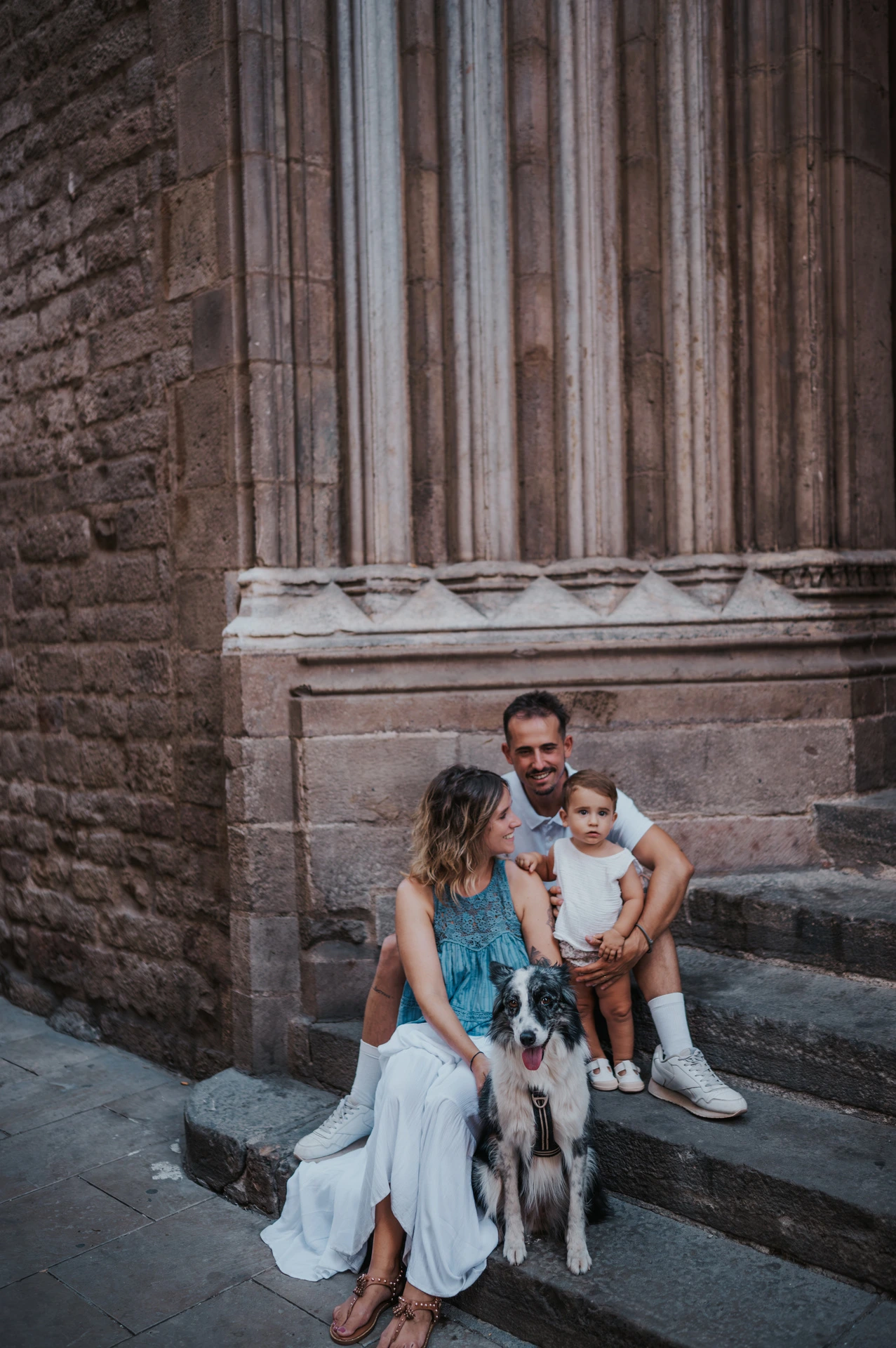 The family sitting on stone steps with their dog in front of them beside a tall carved doorway in El Born during their family photoshoot in Barcelona.