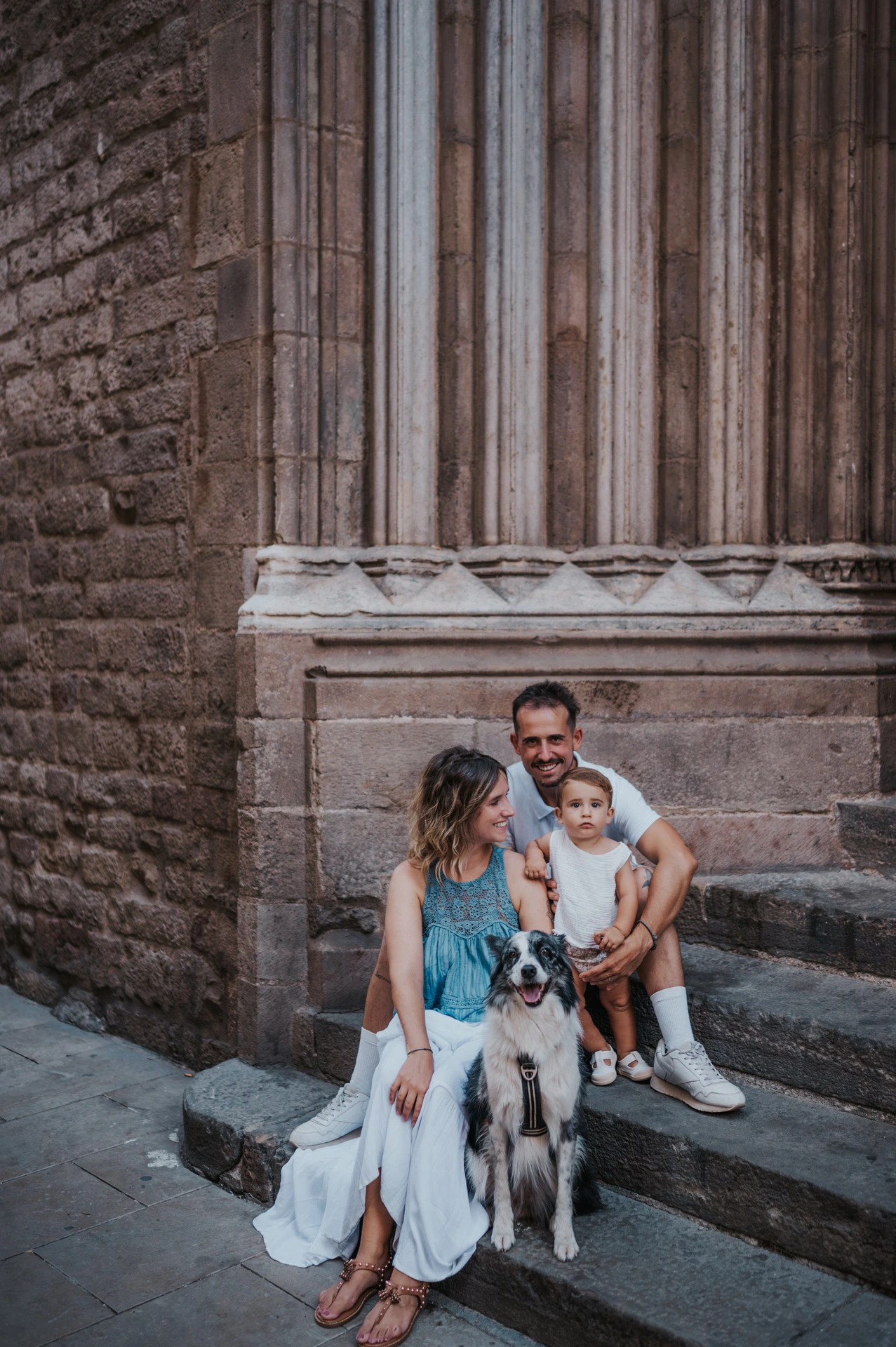 The family sitting on stone steps with their dog in front of them beside a tall carved doorway in El Born during their family photoshoot in Barcelona.