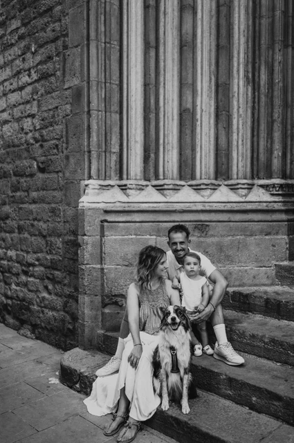 Family portraits with the dog on the stone steps in El Born in black and white | Barcelona family photoshoot Black-and-white photograph: the family sitting on stone steps with their dog in front of them beside a tall carved doorway in El Born during their family photoshoot in Barcelona.