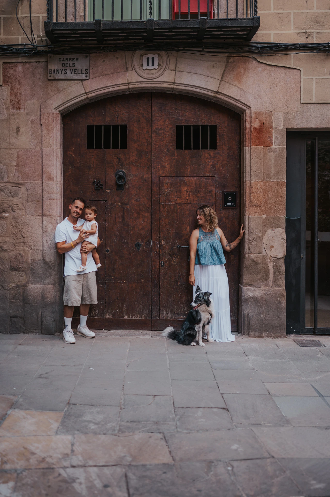 The parents standing apart by broad wooden doors in El Born, with the father holding the toddler and the mother beside their dog during their family photoshoot in Barcelona.