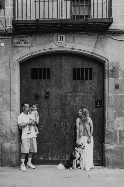 The family and their dog by wooden doors in El Born in black and white | Barcelona family photoshoot Black-and-white photograph: the parents standing apart by broad wooden doors in El Born, with the father holding the toddler and the mother beside their dog during their family photoshoot in Barcelona.
