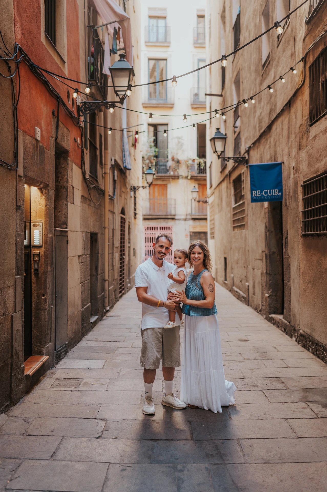 The parents holding their toddler together in a narrow El Born lane lined with balconies during their family photoshoot in Barcelona.