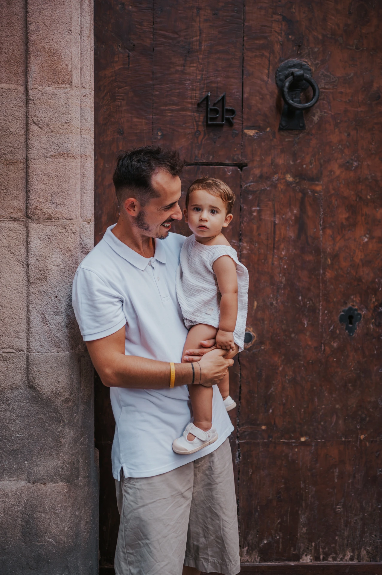 The father holding the toddler beside a weathered wooden doorway in El Born during their family photoshoot in Barcelona.