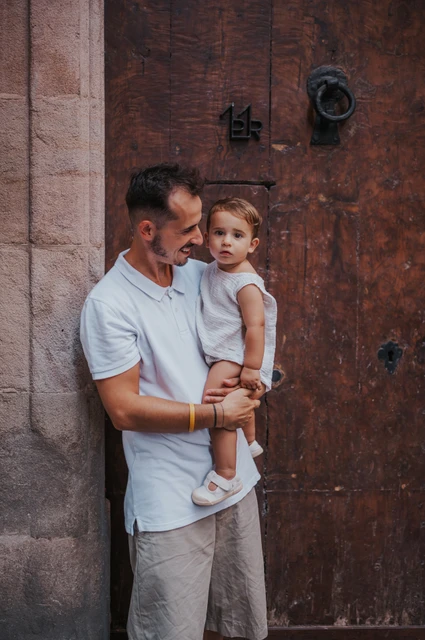 The father holding the toddler beside a weathered wooden doorway in El Born during their family photoshoot in Barcelona.