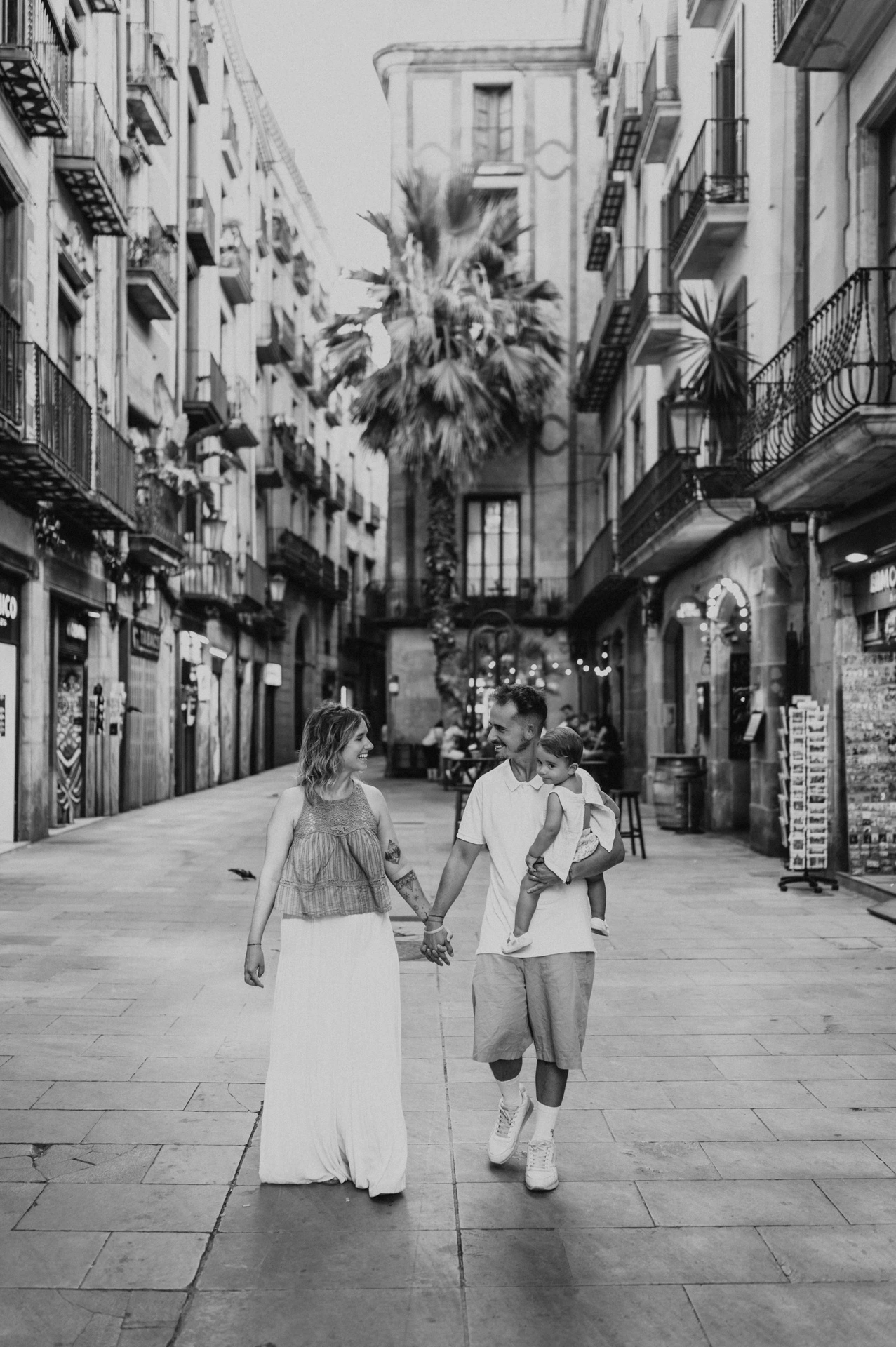Black-and-white photograph: the parents walking hand in hand along a shop-lined street in El Born during their family photoshoot in Barcelona.