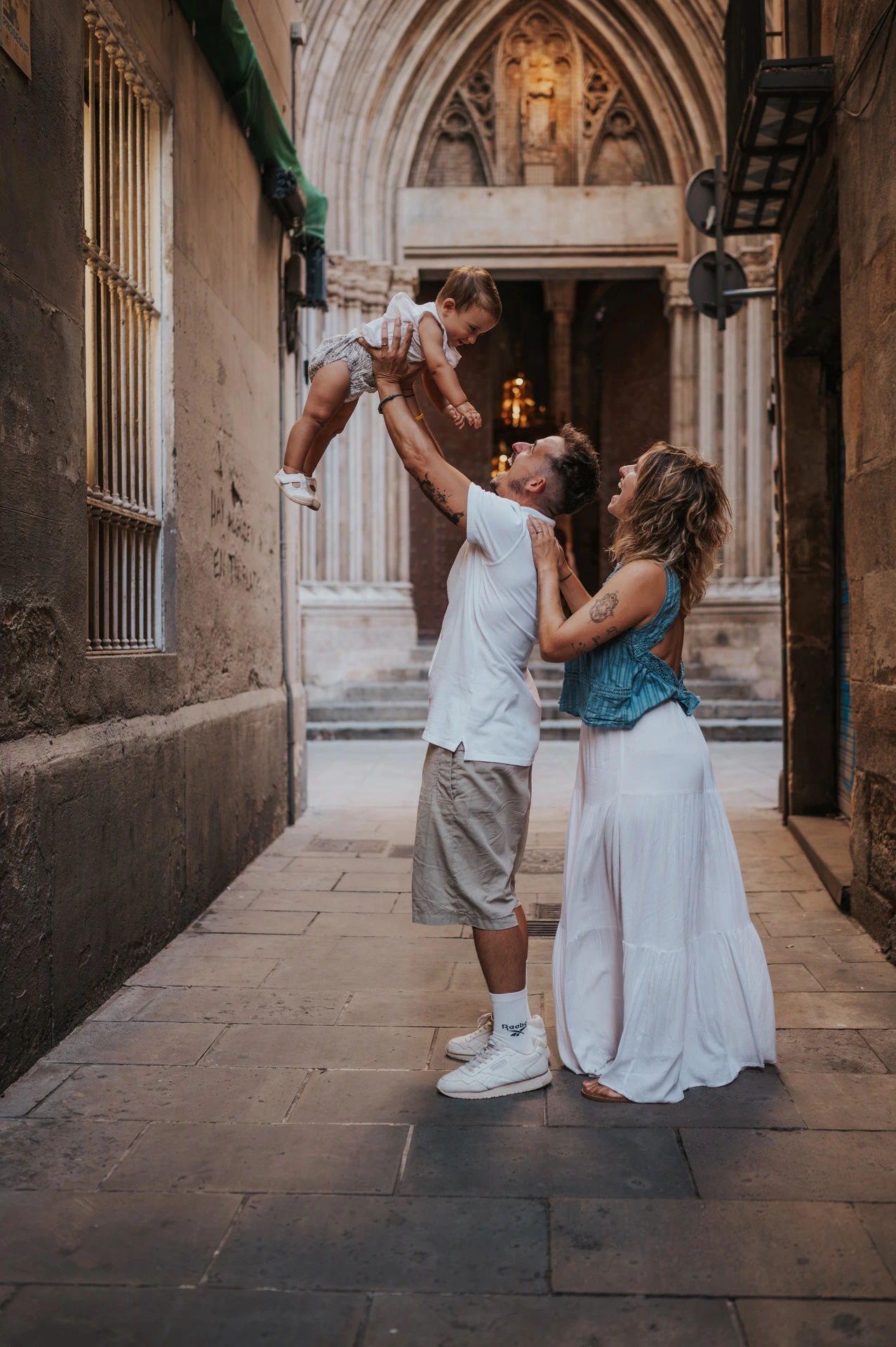 The father lifting the toddler overhead while the mother looks up beside him in a narrow El Born passage during their family photoshoot in Barcelona.