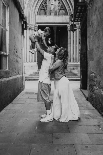 A playful lift beneath an El Born archway in black and white | Barcelona family photoshoot Black-and-white photograph: the father lifting the toddler overhead while the mother looks up beside him in a narrow El Born passage during their family photoshoot in Barcelona.