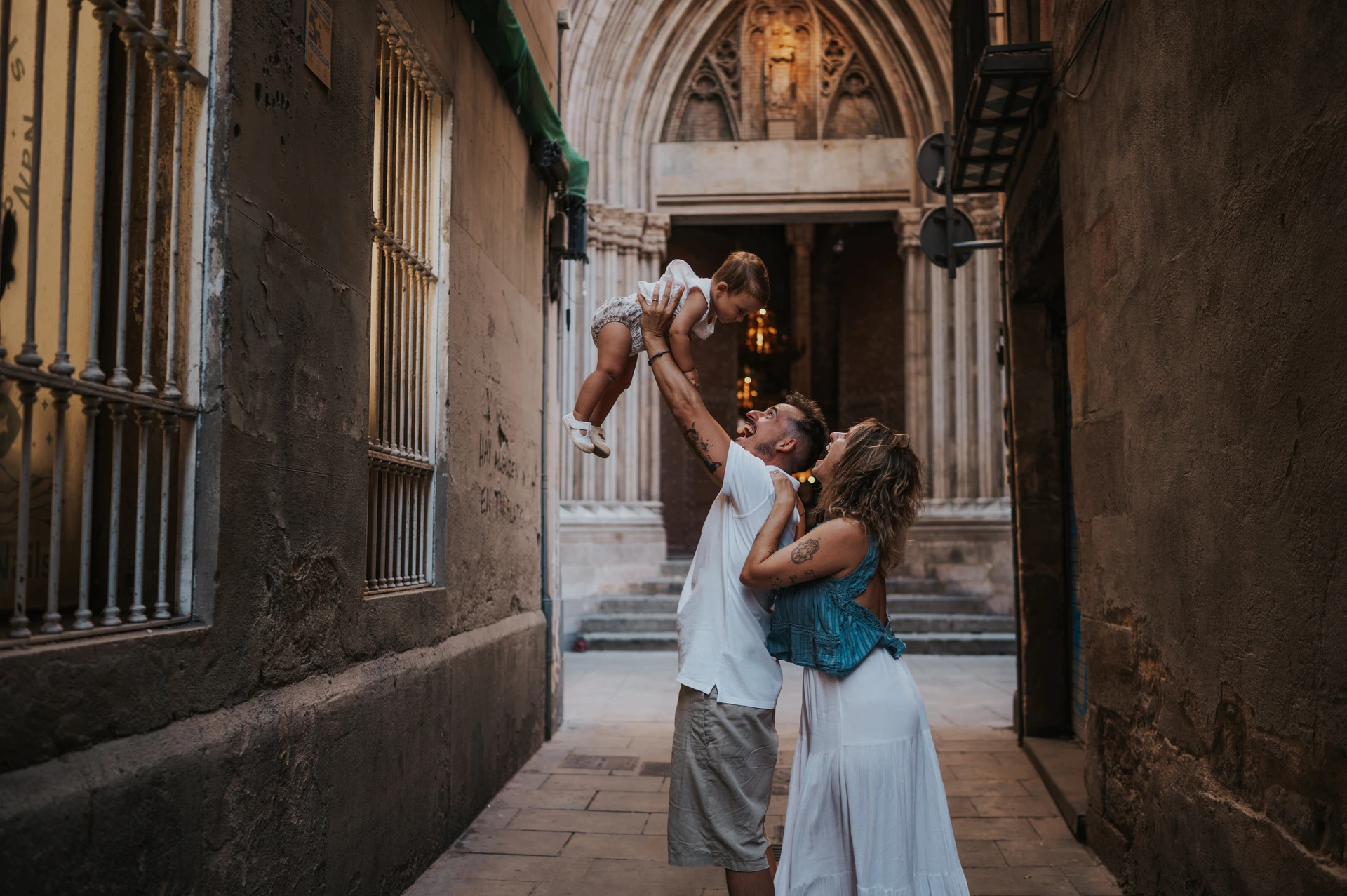 A playful lift beneath an El Born archway | Barcelona family photoshoot The father lifting the toddler overhead while the mother looks up beside him in a narrow El Born passage during their family photoshoot in Barcelona.