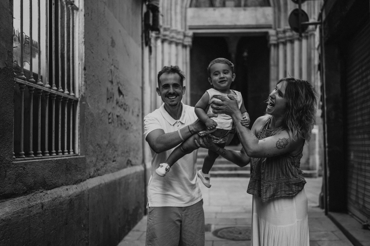 A smiling toddler between the parents in El Born in black and white | Barcelona family photoshoot Black-and-white photograph: the parents holding their smiling toddler between them in an El Born passage during their family photoshoot in Barcelona.