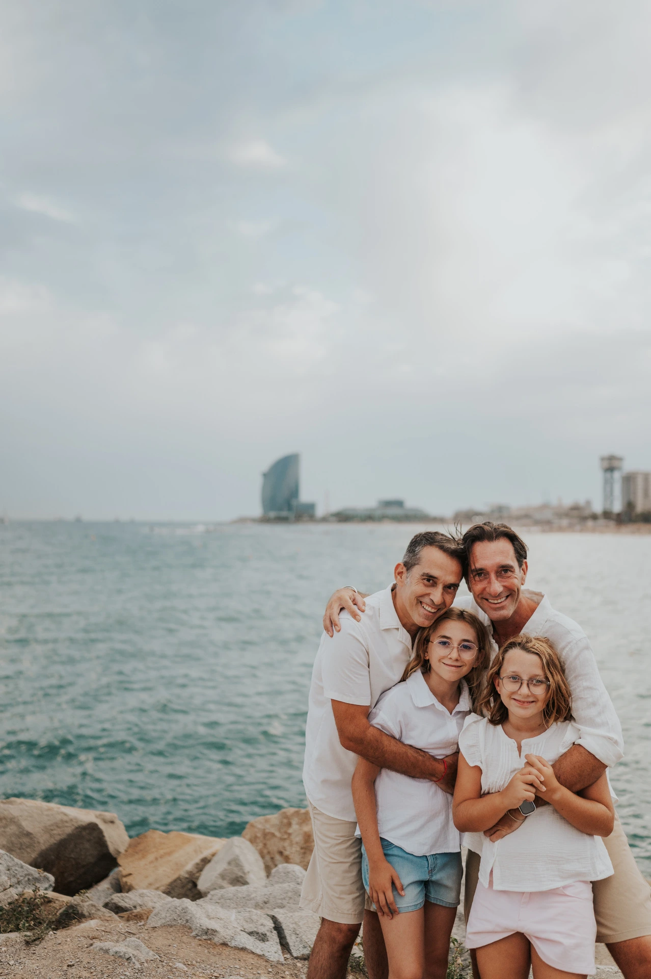 The parents and their two daughters gathered together in a close embrace beside the sea during their family photoshoot in Barcelona.