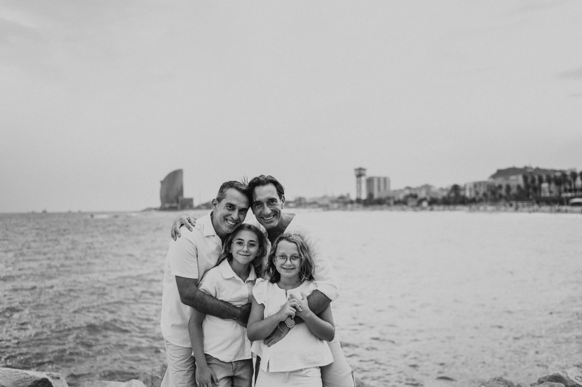 Black-and-white photograph: the parents and their two daughters gathered together in a close embrace beside the sea during their family photoshoot in Barcelona.