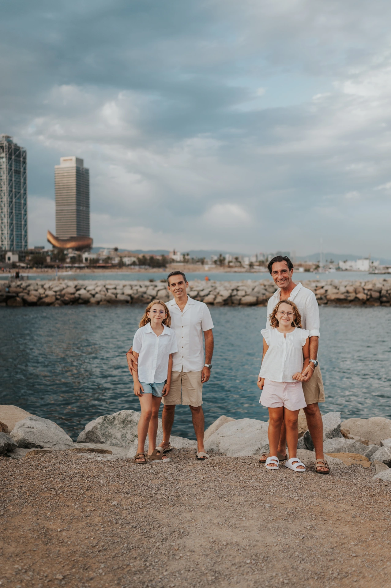 The parents and their two daughters standing together and holding hands beside the sea during their family photoshoot in Barcelona.