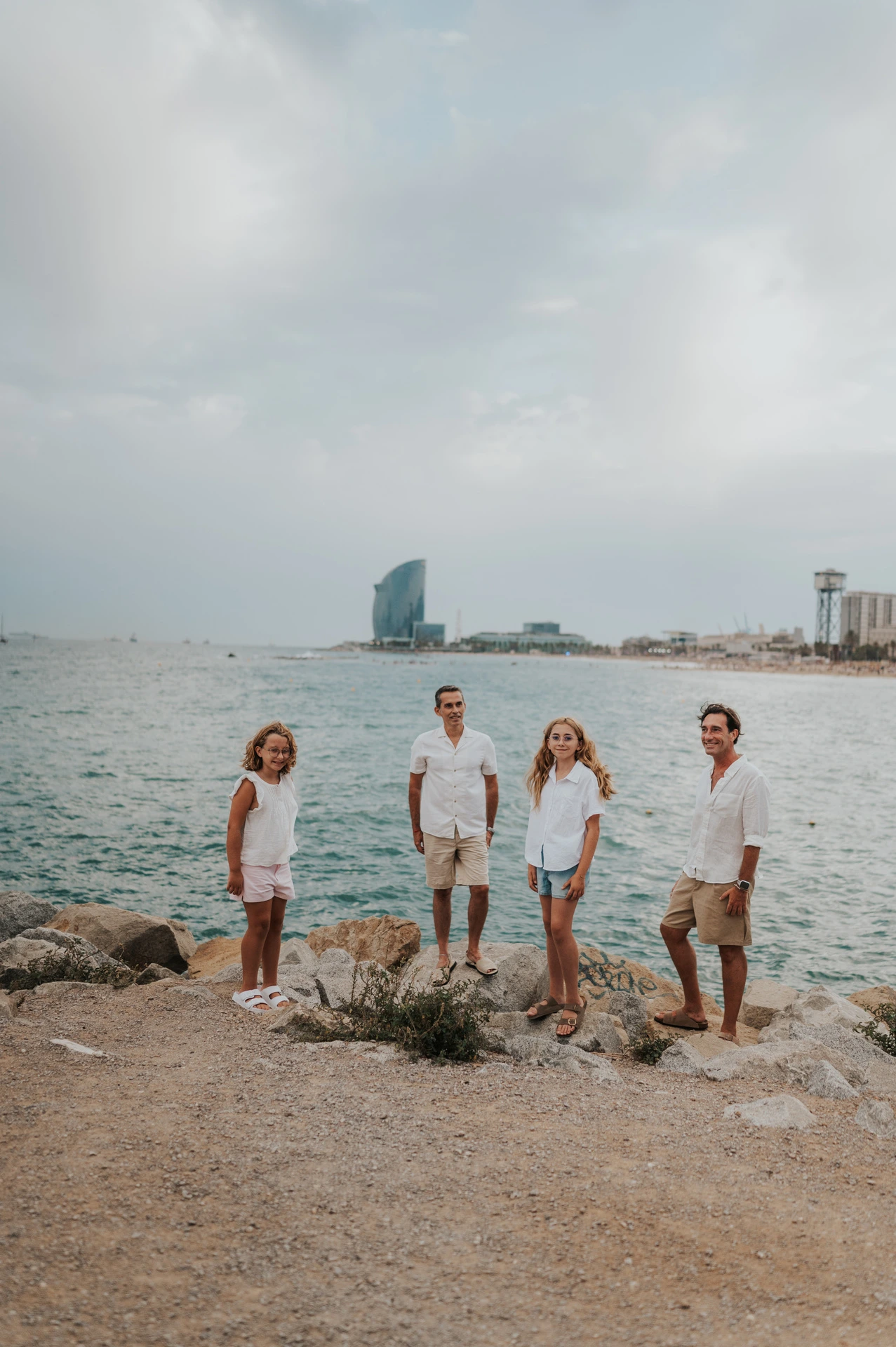 The family of four gathered on the rocky shoreline, with the open sea and W hotel beyond them during their family photoshoot in Barcelona.