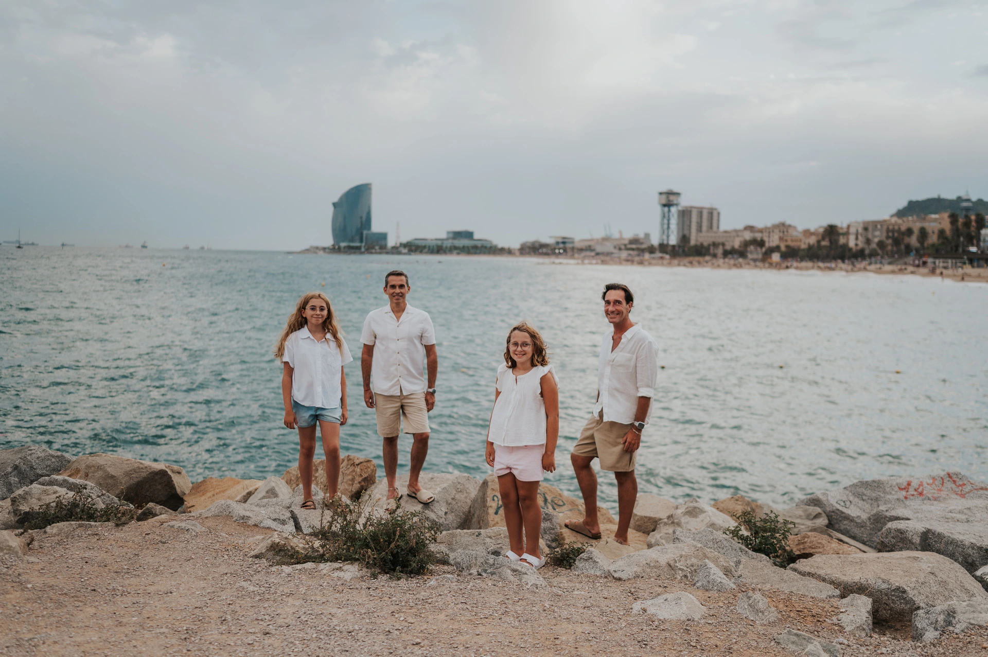 The family of four gathered on the rocky shoreline, with the open sea and W hotel beyond them during their family photoshoot in Barcelona.