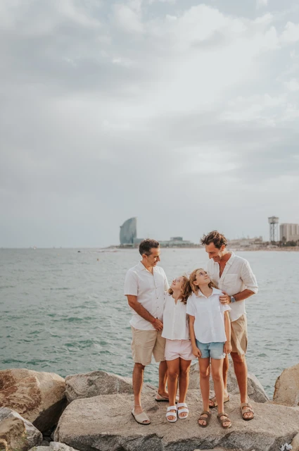 A family portrait on the Barcelona waterfront | Barcelona family photoshoot The family of four standing together beside the waterfront and rocks, with Barcelona’s skyline beyond the sea during their family photoshoot in Barcelona.