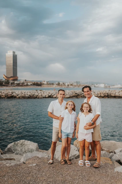 The family of four standing together beside the waterfront and rocks, with Barcelona’s skyline beyond the sea during their family photoshoot in Barcelona.